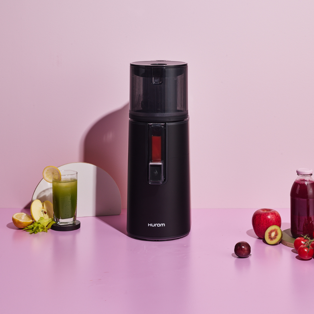A matte black Hurom H400 Easy Clean Slow Juicer, facing the front, with a cup of juice beside and fruits beside it.
