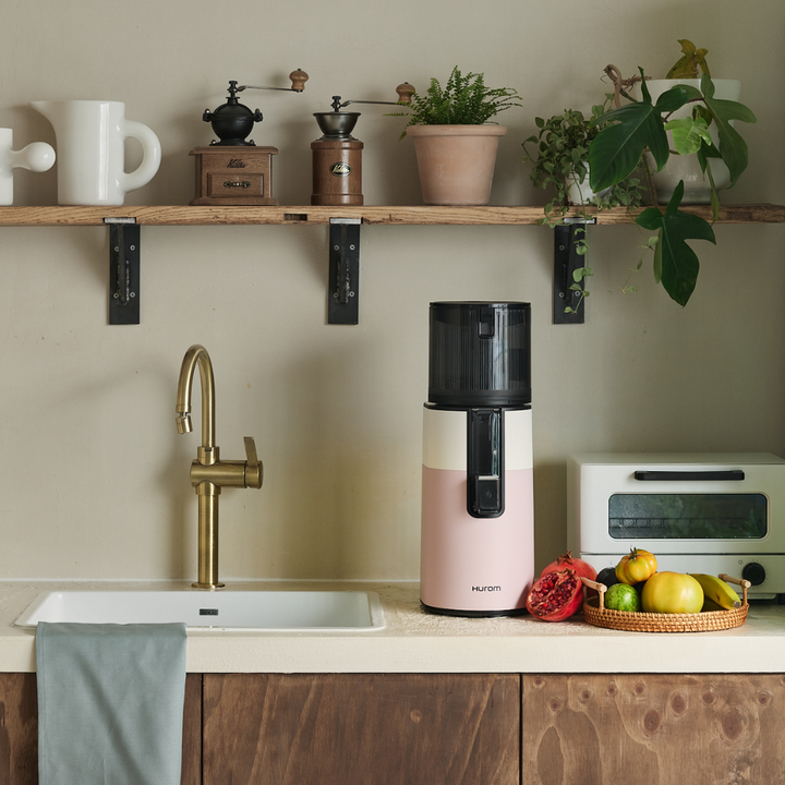 A white pink Hurom H400 Easy Clean Slow Juicer on a kitchen top, facing the front, with a kitchen sink and an oven beside.