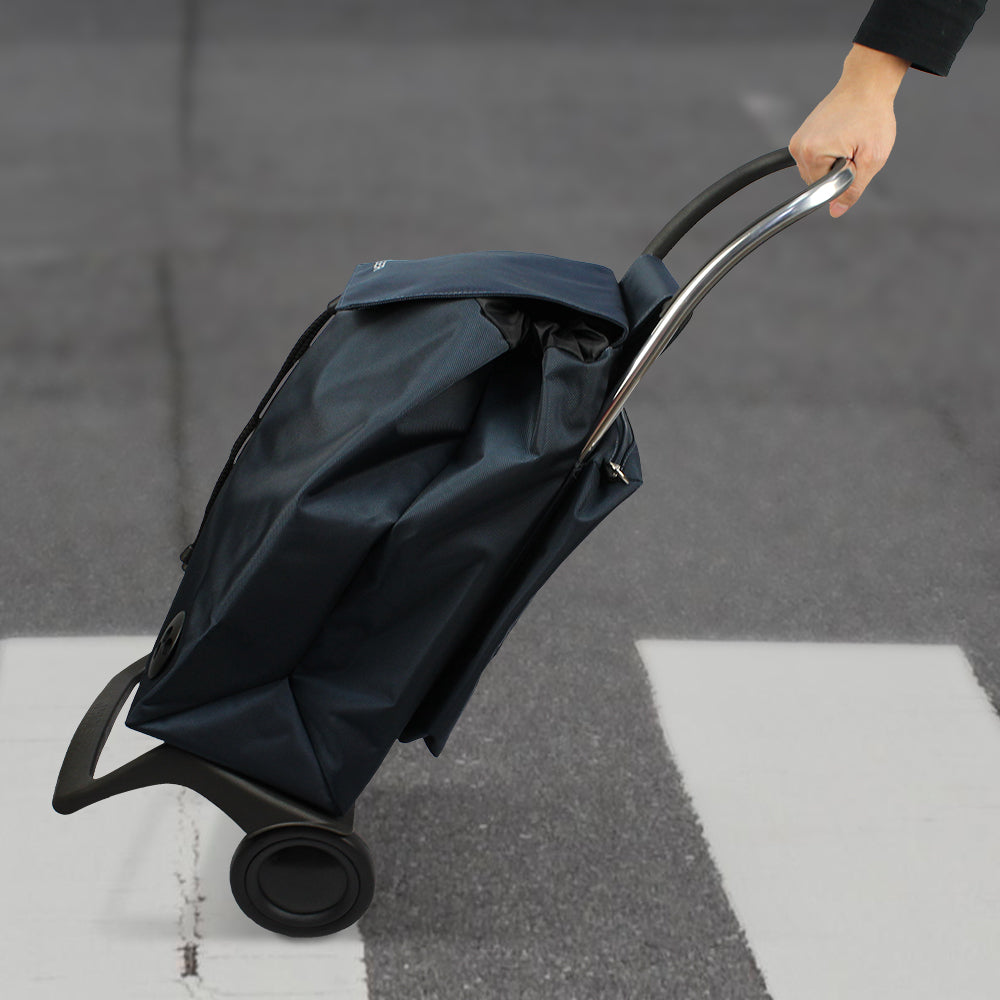 A blue Rolser Baby Joy Market & Shopping Trolley with a thermo bag, rolling on the ground with a man pulling on it.